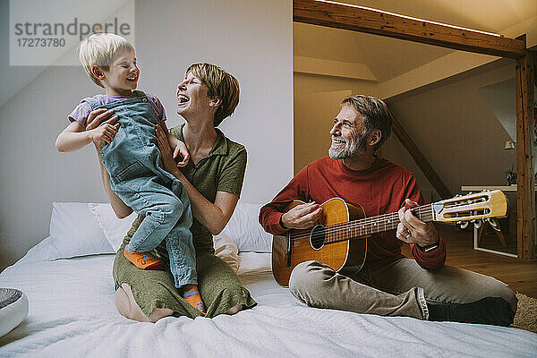 Vater spielt Gitarre  während Mutter und Sohn auf dem Bett sitzend im Schlafzimmer zu Hause Musik genießen
