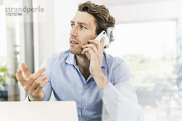 Mittlerer erwachsener Mann  der mit seinem Smartphone spricht und wegschaut  während er im Büro sitzt