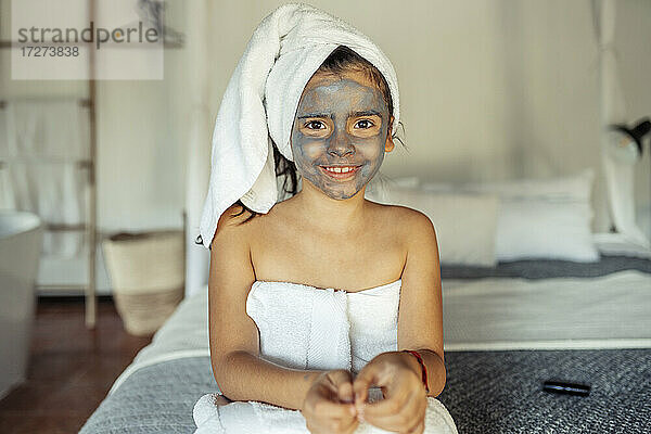 Lächelndes Mädchen  bedeckt mit Handtuch und Gesichtsmaske  sitzt auf dem Bett zu Hause
