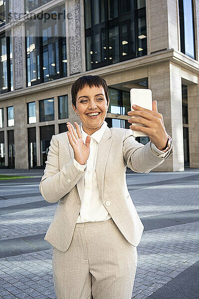 Frau winkt mit der Hand zu einem Videoanruf auf einem Mobiltelefon  während sie vor einem modernen Bürogebäude steht