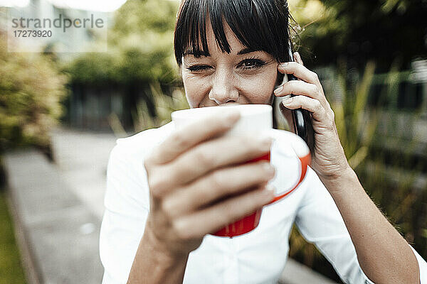 Lächelnde Geschäftsfrau  die eine Kaffeetasse hält  während sie mit einem Mobiltelefon spricht