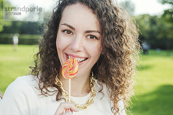 Schöne glückliche junge Frau mit lockigem braunem Haar isst Lutscher im Park