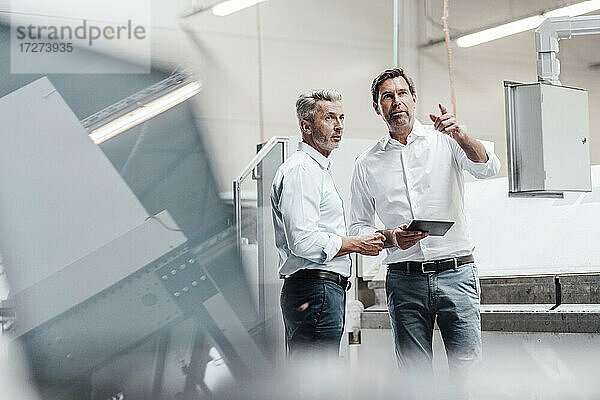 Männlicher Ingenieur mit digitalem Tablet  der auf eine Tafel zeigt  während er mit einem Kollegen in der Fertigungsindustrie diskutiert