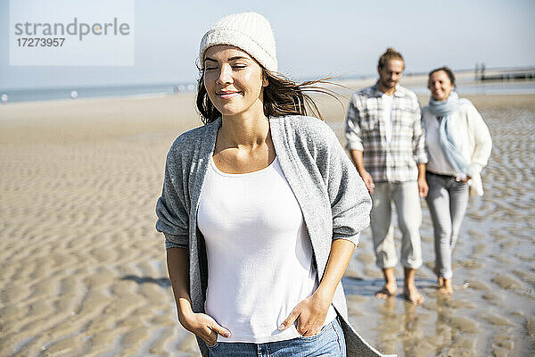 Lächelnde Frau  die mit ihrem Freund und ihrer Mutter im Hintergrund am Strand spazieren geht