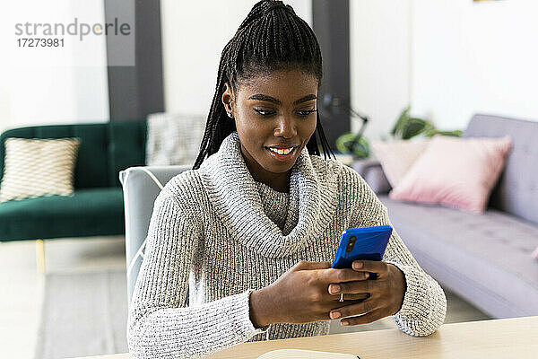 Lächelnde junge Frau  die eine SMS auf ihrem Smartphone schreibt  während sie zu Hause im Wohnzimmer sitzt