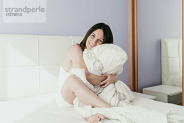 Glückliche schöne Frau umarmt Decke sitzt auf dem Bett zu Hause