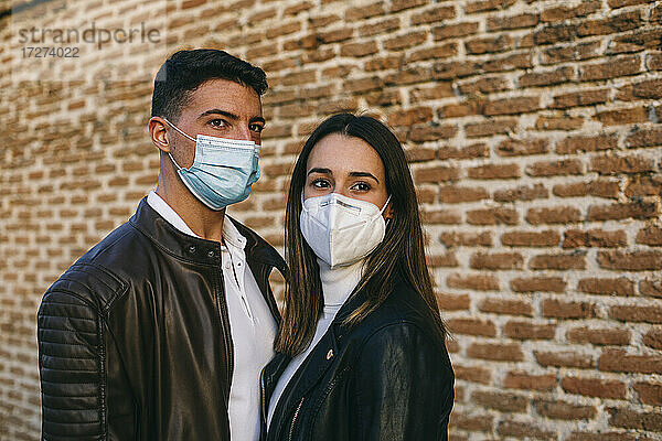 Junges Paar  das eine Gesichtsmaske trägt und wegschaut  während es zusammen an einer Mauer steht