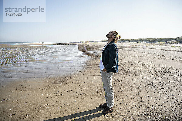 Junger Mann mit Händen in den Taschen am Strand stehend gegen den klaren Himmel