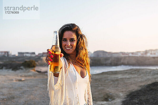 Lächelnde Frau zeigt kleine Weißweinflasche am Strand bei Sonnenuntergang