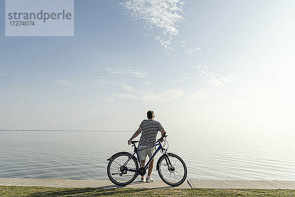 Mann steht mit Fahrrad und schaut auf das Meer vor blauem Himmel