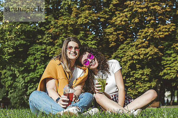 Lächelnde Freundinnen sitzen mit frischer Limonade im Gras im Park an einem sonnigen Tag