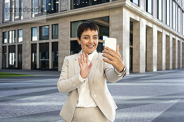 Geschäftsfrau winkt mit der Hand zum Videoanruf  während sie vor einem modernen Bürogebäude steht