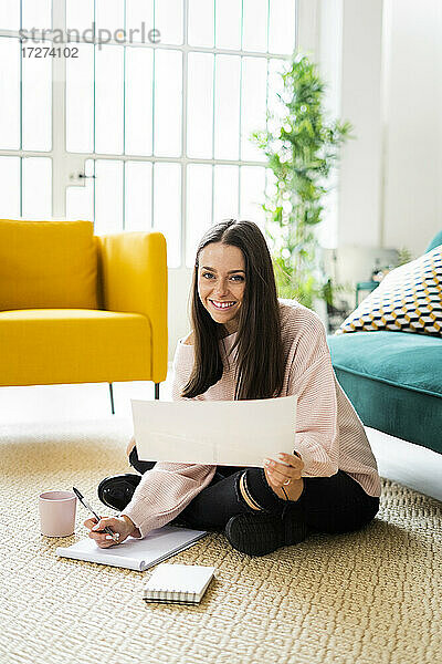 Glückliche schöne junge Frau sitzt mit Notizblöcken und Kaffeetasse auf Teppich vor Sofa zu Hause
