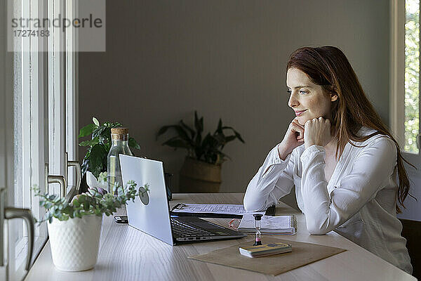 Weiblicher Arzt mit Hand am Kinn  der zu Hause sitzend einen Videoanruf auf dem Laptop hört