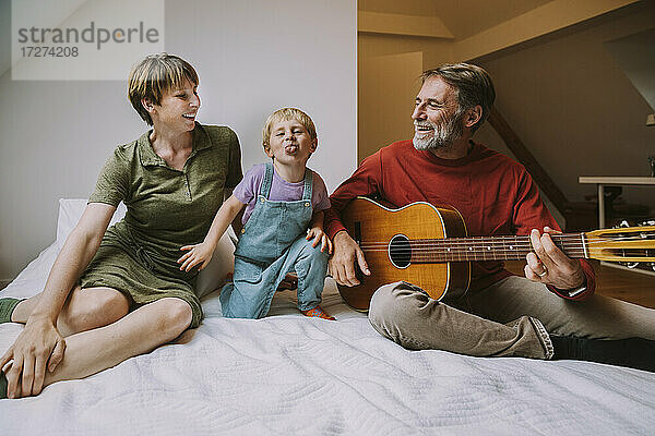 Sohn streckt die Zunge heraus  während der Vater Gitarre spielt und die Mutter neben ihm auf dem Bett sitzt