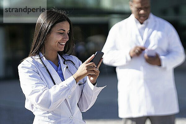 Lächelnde Frau  die ein Mobiltelefon benutzt  während sie mit einem Arzt im Hintergrund vor einem Krankenhaus steht