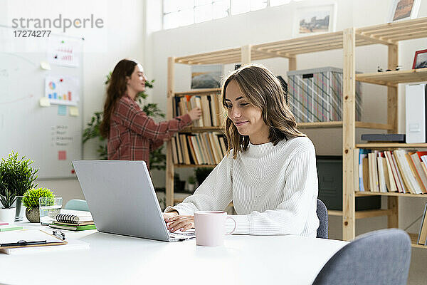 Lächelnde Geschäftsfrau  die an einem Laptop arbeitet  während ein Kollege im Hintergrund im Büro steht