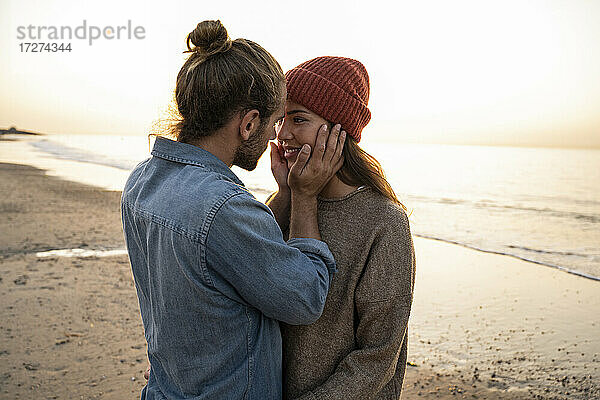 Romantischer junger Mann  der das Gesicht seiner Freundin berührt  während er bei Sonnenuntergang am Strand steht