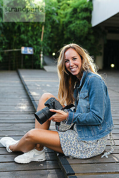 Lächelnde schöne blonde Frau  die eine Digitalkamera hält  während sie auf einem Fußweg sitzt