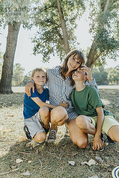 Glückliche Mutter  die ihre Söhne umarmt  während sie in einem öffentlichen Park an einem sonnigen Tag kniet