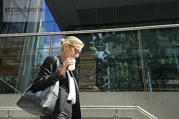 Lächelnde Frau  die eine Kaffeetasse in der Hand hält  während sie auf einer Treppe gegen ein Gebäude läuft