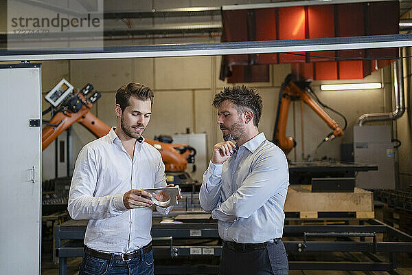 Geschäftsmann mit Metallgegenstand  der mit einem Kollegen diskutiert  während er in einer Fabrik vor einer Robotermaschine steht
