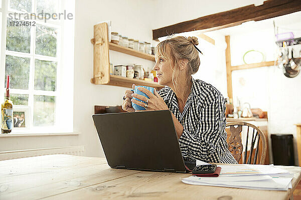 Ältere Frau  die wegschaut  während sie Kaffee trinkt und mit einem Laptop in der Küche zu Hause sitzt