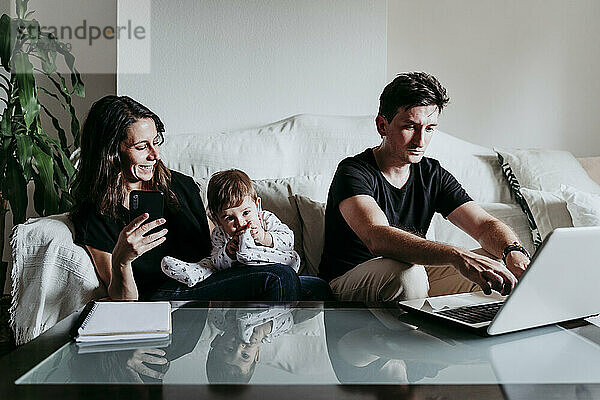 Lächelnde Mutter  die ein Smartphone benutzt  während der Vater am Laptop arbeitet und der Sohn zu Hause sitzt