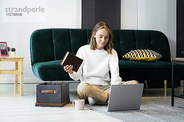 Geschäftsfrau mit Buch  die einen Laptop benutzt  während sie bei einer Kiste auf dem Boden im Büro sitzt