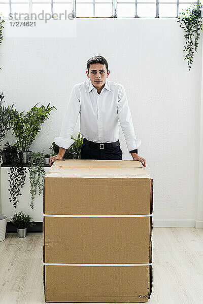 Selbstbewusster männlicher Fachmann  der sich auf einen Karton an einer weißen Wand am Arbeitsplatz stützt