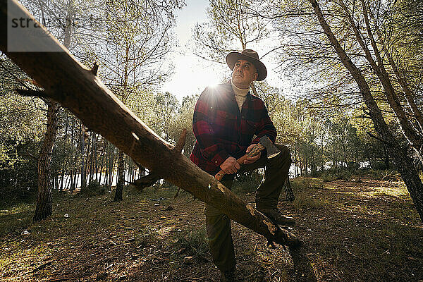 Buschmann mit Axt am Baumstamm stehend und wegschauend im Wald
