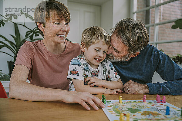 Vater kuschelt mit seinem Sohn  während die Mutter daneben sitzt und zu Hause ein Brettspiel spielt