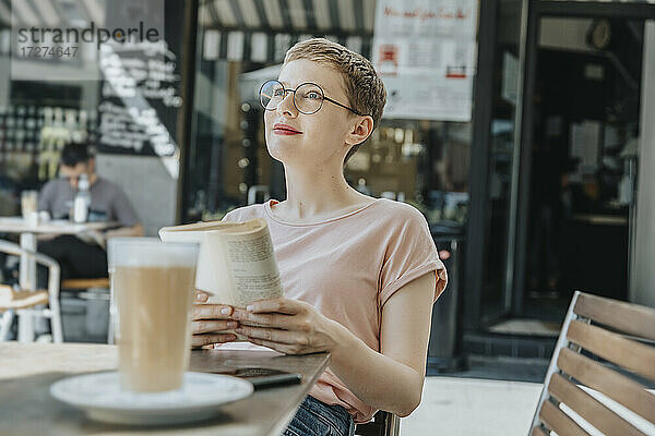 Mittlere erwachsene Frau  die mit einem Buch wegschaut  sitzt in einem Straßencafé an einem sonnigen Tag