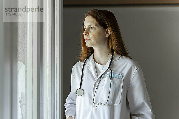 Nachdenkliche junge Ärztin  die durch das Fenster schaut  während sie in ihrem Büro zu Hause steht