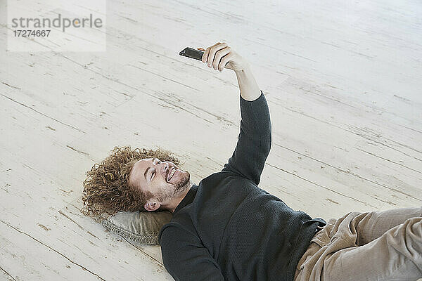Lächelnder rothaariger Mann  der ein Selfie mit seinem Smartphone macht  während er zu Hause auf dem Boden liegt