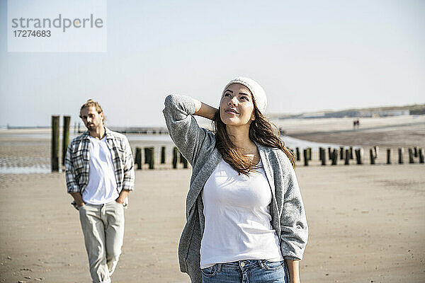 Nachdenkliche junge Frau schaut auf  während ein Mann im Hintergrund am Strand spazieren geht