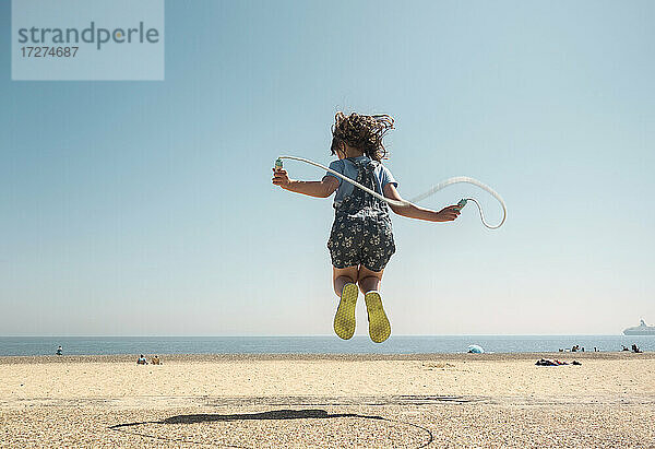 Mädchen spielt mit Springseil und springt am Strand gegen den klaren Himmel