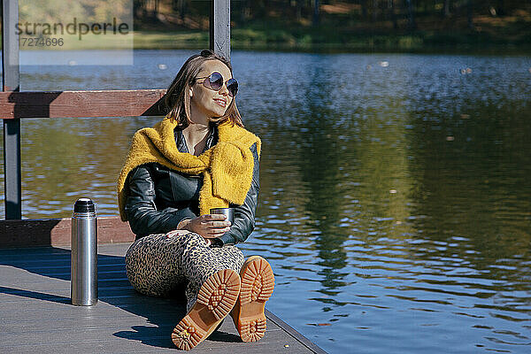 Lächelnde Frau  die Kaffee trinkt  während sie sich am Wochenende auf dem Pier über dem See entspannt