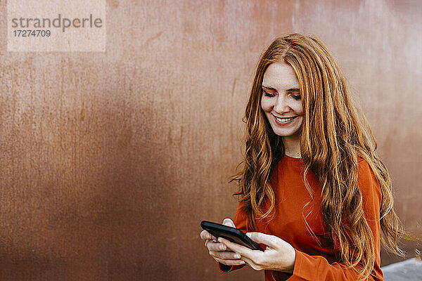 Rothaarige junge Frau lächelt  während sie eine Textnachricht über ihr Mobiltelefon an die Wand schreibt