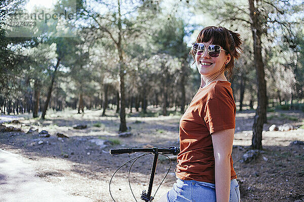 Lächelnde junge Frau mit Sonnenbrille und Fahrrad auf dem Lande am Wochenende