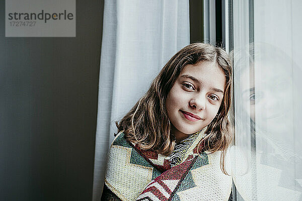 Lächelndes  in eine Decke gehülltes Mädchen  das zu Hause am Fenster sitzt
