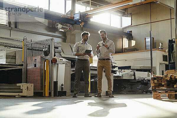 Geschäftsmann mit digitalem Tablet  der neben einem Kollegen steht  der ein Metallobjekt in einer Fabrik überprüft