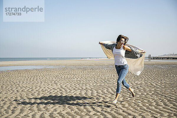 Unbeschwerte junge Frau  die mit einer Decke in der Hand am Strand gegen den klaren Himmel läuft