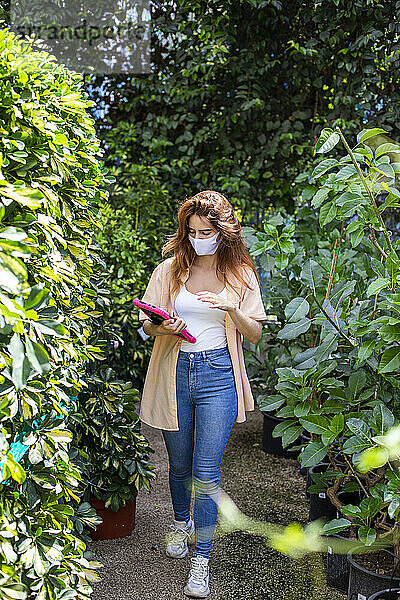 Geschäftsfrau mit Gesichtsmaske  die ein digitales Tablet benutzt  während sie in einem Gartencenter spazieren geht