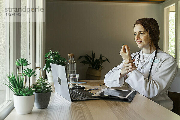 Junge Ärztin  die das Handgelenk hält  während sie während einer Online-Konsultation im Büro zu Hause etwas erklärt