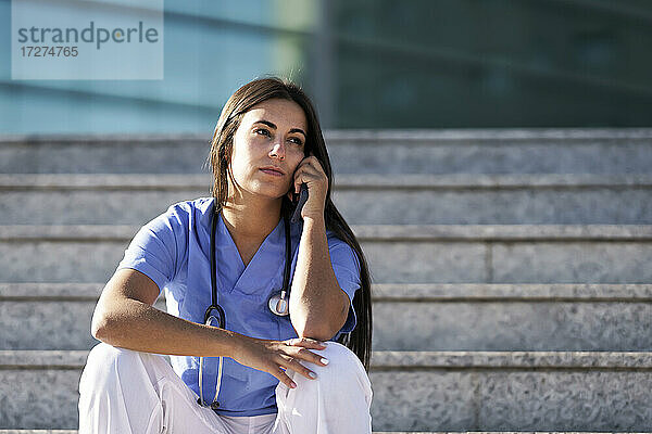 Junger Arzt  der mit seinem Handy telefoniert  während er auf der Treppe vor dem Krankenhaus sitzt