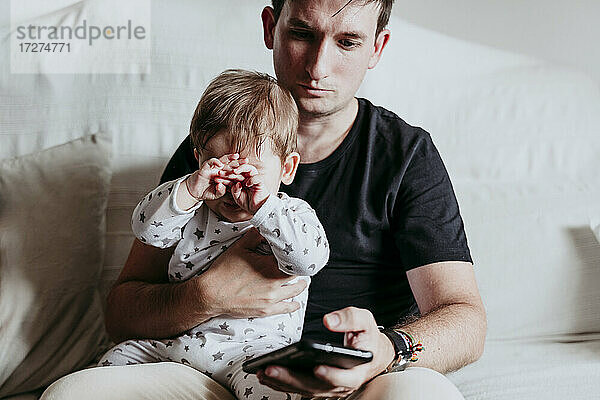 Vater benutzt sein Smartphone  während er mit seinem Sohn zu Hause auf dem Sofa sitzt