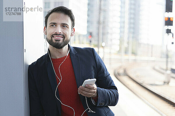 Lächelnder Unternehmer mit In-Ear-Kopfhörern wartet auf dem Bahnsteig