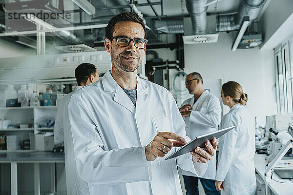Lächelnder männlicher Wissenschaftler  der ein digitales Tablet benutzt  während er mit einem Mitarbeiter im Hintergrund in einem Labor steht
