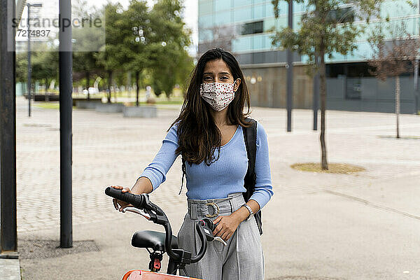 Schöne Frau mit Schutzmaske  die während der COVID-19-Pandemie mit dem Fahrrad in der Stadt steht